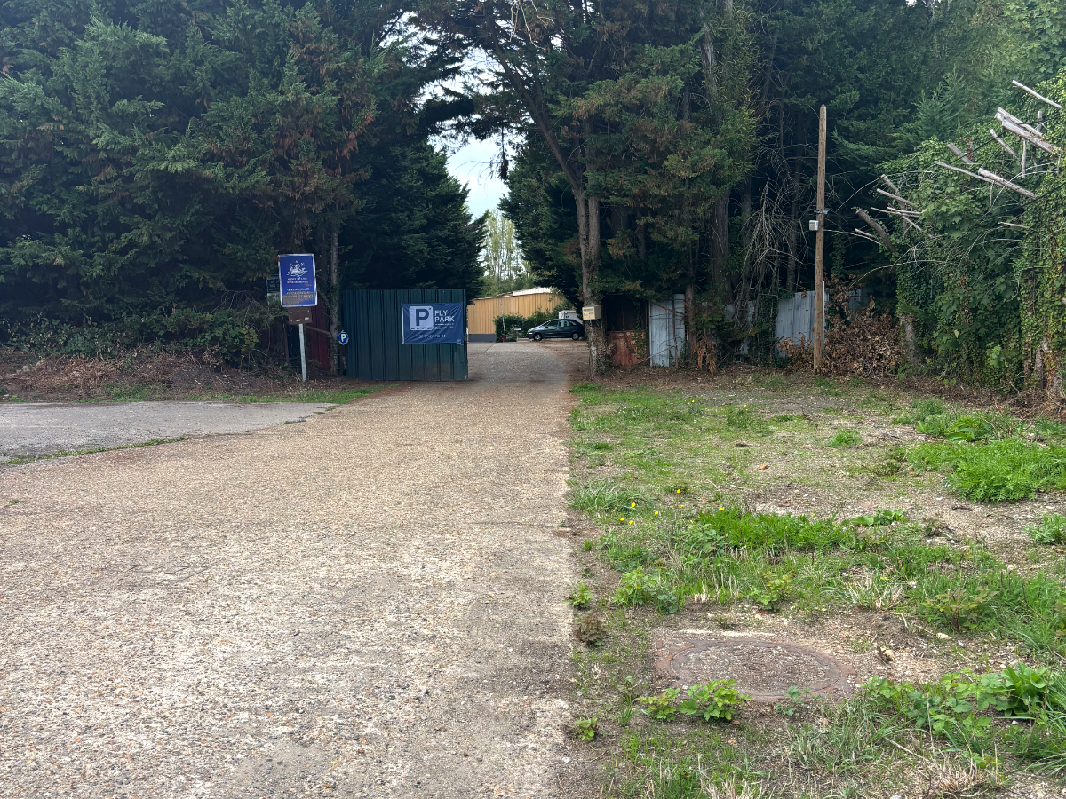 Allée d'entrée gravillonnée du parking Fly Park, vue depuis la route, avec la signalétique bleue